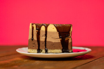 Chocolate syrup is poured over a chocolate cake that on a plate on a pink background. 