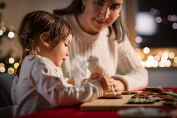 family, cooking and winter holidays concept - happy mother and baby daughter with icing in baking bag decorating gingerbread cookies at home on christmas