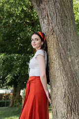 Pretty model in blouse and skirt standing near tree in park.