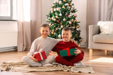 christmas, winter holidays and childhood concept - happy girl and boy in pajamas with gifts sitting on floor and hugging at home