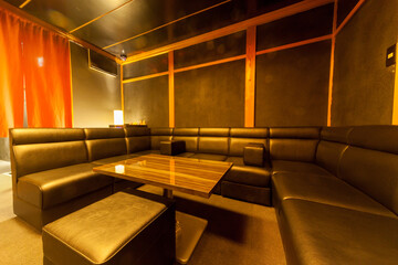 繁華街にある落ち着いた雰囲気のスナックやバーの店内。L字型の黒いレザーソファーと木目調のテーブルが並ぶ、高級感のあるカラオケ個室やラウンジの内観写真。テキストを入れるスペースあり