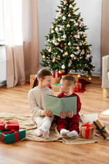 christmas, winter holidays and childhood concept - happy girl and boy in pajamas reading book together sitting on floor in front of each other at home
