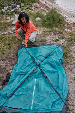 Vertical high angle view of young adult Hispanic woman setting up camp at lakeside inserting poles into flaps in tent