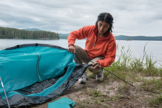 Young adult Hispanic woman spending weekend travelling at lakeside setting up tent inserting poles into flaps