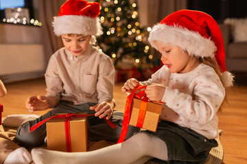 Obraz na płótnie Canvas christmas, winter holidays and childhood concept - happy little girl and boy opening gifts sitting on floor at home