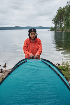 Vertical shot of young adult Hispanic woman spending weekend at lake shore setting up camp tent fixing poles on top