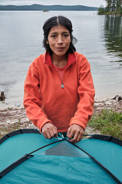 Vertical medium portrait of young adult Hispanic traveller standing at lake shore fixing tent poles on top looking at camera