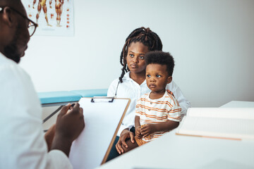 A sweet little African boy sits up on the exam table at the doctors, as his male doctor listens to...