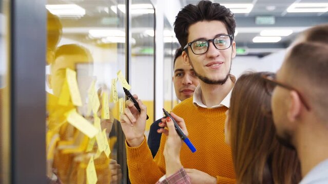 Business team brainstorm writing ideas on paper sticky notes and post it on glass wall. Startup coworkers discuss a strategy of work plan for product development at scrum meeting in corporate office.