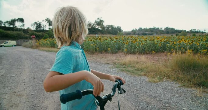 Child with bicycle looks out into the distance at the horizon beyond sunflower field at countryside. Childhood on rural environment. Children on summer vacation in village