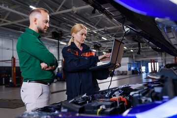 A young female car mechanic holds a diagnostic laptop and shows the manager the identified errors against the background of a large service center