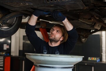 Fototapeta na wymiar Car maintenance. A male car mechanic in uniform changes the oil in the engine while standing under the car on the lift