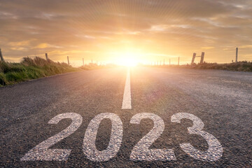 Sign 2023 on a small straight asphalt road at sunrise. New year plans and resolution. Warm color tone. Sun flare and glow.