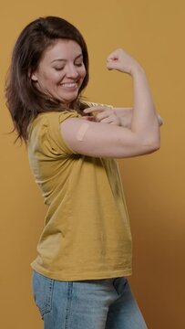 Portrait Of Smiling Woman Lifting Sleeve And Showing Band Aid After Covid Or Flu Vaccine Flexing Arm Biceps In Studio. Proud Person Revealing Bandage Covering Immunization Spot For Medical Awareness.