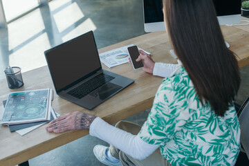 Queer designer using smartphone with blank screen near papers in office.