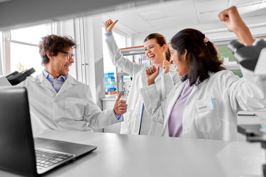 science, work and people concept - international group of happy smiling scientists with laptop celebrating success in laboratory