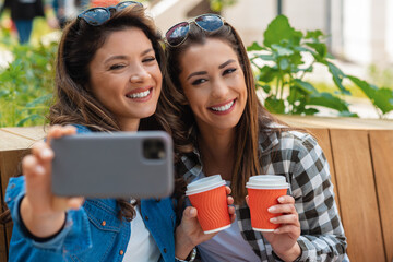 Two best friend taking break from shopping. Two woman sitting outdoors drinking coffee and taking photo with phone