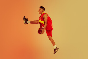 Fototapeta na wymiar Portrait of young man, professional basketball player playing isolated over orange studio background in neon light. Ball possession