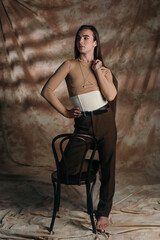 Trendy nonbinary person in corset holding jacket near chair on abstract brown background.