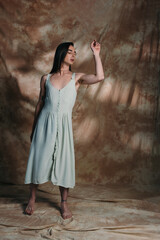 Barefoot nonbinary person in light blue sundress posing on abstract brown background.