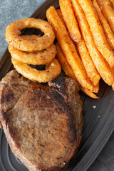 Rump steak dinner, with sweet potato fries and onion rings, on a black oval plate.  On a dark stone background