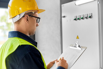 construction business and building concept - male electrician or worker in helmet and safety west with papers on clipboard and pencil at electric board