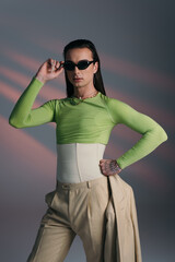 Brunette nonbinary model in corset and sunglasses holding jacket on abstract background.