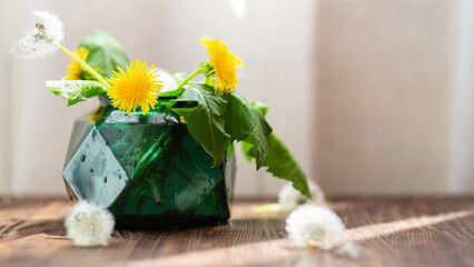 A bouquet of summer dandelions in a green round vase on a wooden table in the rays of the sun
