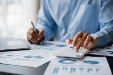 Businessman is using a calculator to calculate company financial figures from earnings papers, a businessman sitting in his office where the company financial chart is placed. Banner with copy space.