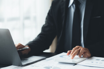 Businessman is using a calculator to calculate company financial figures from earnings papers, a businessman sitting in his office where the company financial chart is placed. Banner with copy space.