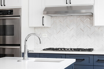 A luxury kitchen sink detail shot with marble countertops, herringbone tile backsplash, and white and blue cabinets.
