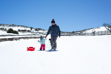 Skier father and his little daughter walking on an empty ski slope with a red sledge. Winter activities in family. - obrazy, fototapety, plakaty