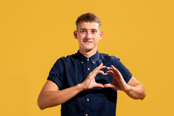 Portrait of curious charming guy showing heart symbol on yellow background