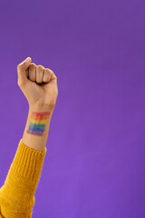 Close up of biracial man raising fist with lgbt flag on arm on purple background