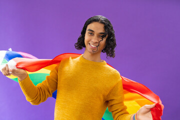 Portrait of happy biracial man holding lgbt rainbow flag and smiling on purple background