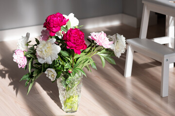 Ornate bouquet of various peonies in transparent vase located on the floor. White chair in background. Blurred room in background.