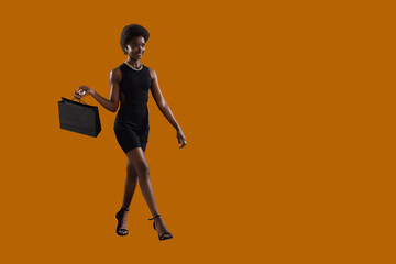 Fototapeta na wymiar African-american woman full height walking with black shopping bag on brown background. Sale and discounts concept.