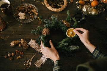 Millennial woman making Christmas wreath using natural pine branches and festive decorations in workshop.