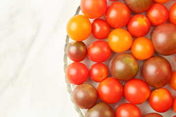 Top down view of a variety of fresh, organic, different color and size cherry tomatoes in a flat glass plate on a white marble kitchen board. Copy space