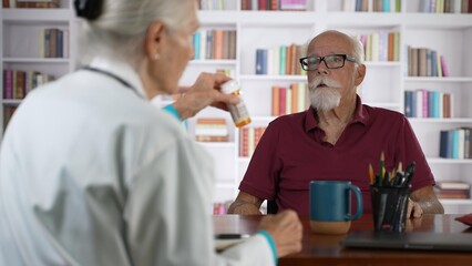 Obraz premium Mature senior male doctor discussing treatment with prescription drug to female patient in modern clinic with books background.