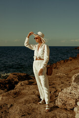 Elegant fashionable woman wearing white linen suit, straw hat, sunglasses, leather sandals, with...