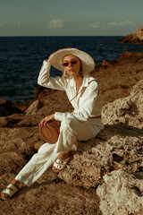 Elegant fashionable woman wearing white linen suit, straw hat, sunglasses, leather sandals, with round wicker bag, posing on rocks near sea. Outdoor summer fashion full-length  portrait 