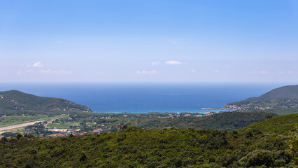 View from wooded mountains to blue waters of gulf and beach near town of Procchia on a clear sunny day. Province of Livorno, Island of Elba, Italy