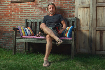 Blond man in black t-shirt, green shorts and sandals sits on a bench in backyard.