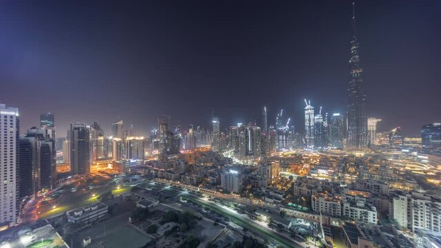 Dubai Downtown during all night timelapse panorama with tallest skyscraper and other illuminated towers view from the top in Dubai, United Arab Emirates. Moon on the sky and lights turning off