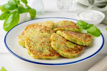 Zucchini fritters with cheese, garlic and herbs. Vegetarian zucchini pancakes, served with basil and sour cream on a white wooden background. Selective focus.