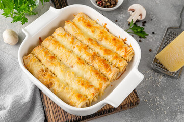 Pancakes or thin crepes stuffed with chicken and mushrooms baked with cheese in a baking dish on a gray concrete background
