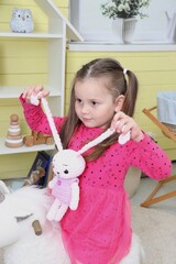 A little girl pulls the ears of a naughty pink hare in the children's room. A little girl in a pink dress plays alone in the room. Knitted handmade toys in the hands of a child. A child on a unicorn
