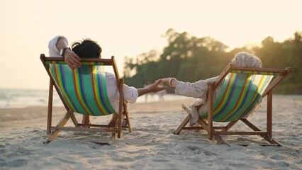 Happy Asian family senior couple enjoy outdoor lifestyle on summer beach holiday vacation at the sea. Elderly retired man and woman relaxing on beach chair and holding hands together at summer sunset - Powered by Adobe