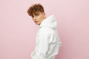 a handsome, pleasant young man stands on a pink background in a white hoodie and looks at the...